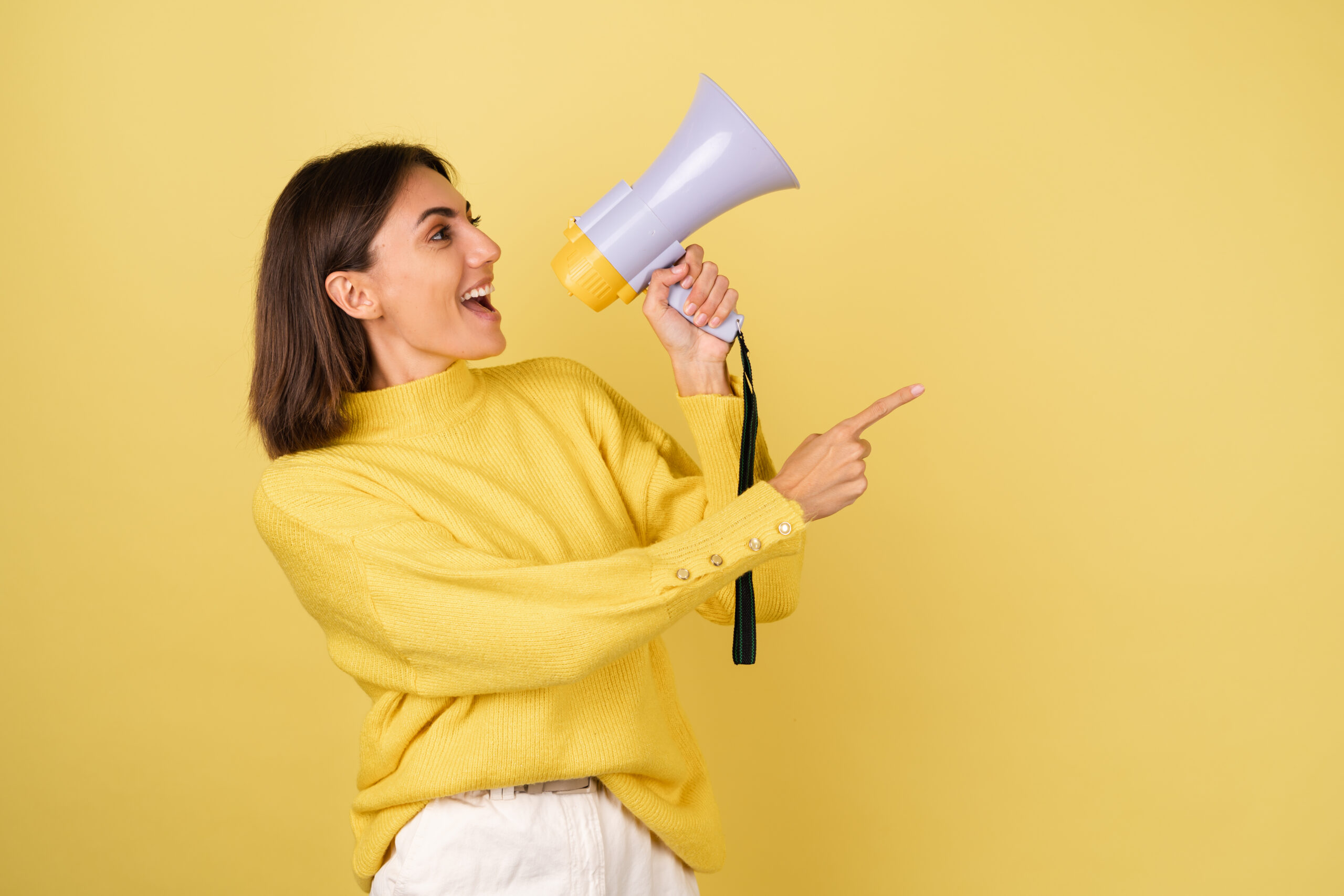 Mujer joven sonriente vestida de amarillo, sosteniendo un megáfono y señalando con entusiasmo hacia la derecha sobre fondo amarillo